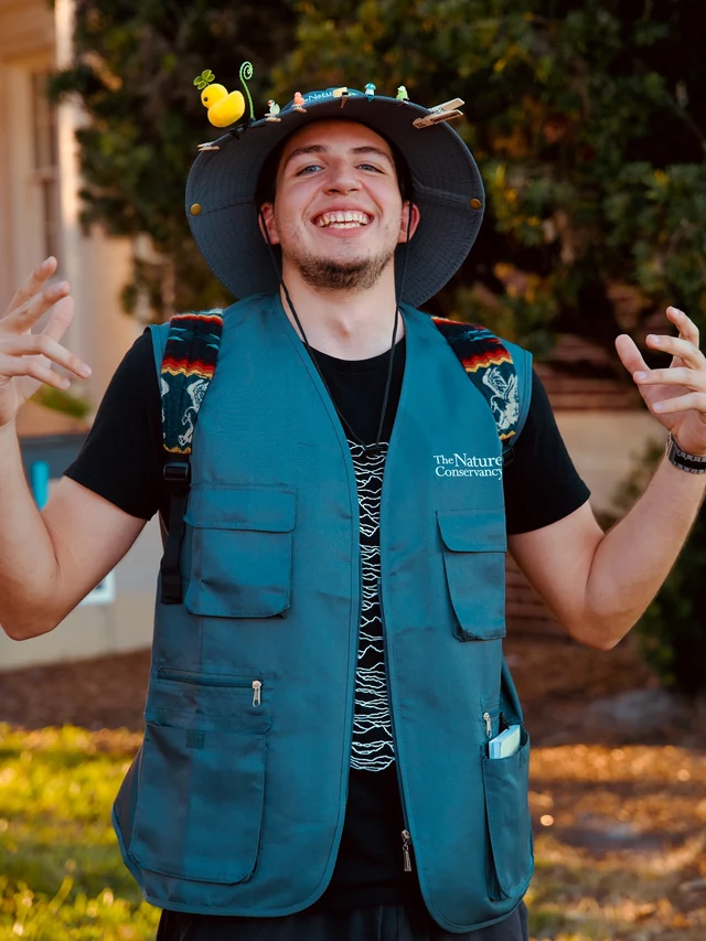 A smiling man wearing a blue nature conservancy vest and a hat decorated with tiny ducks