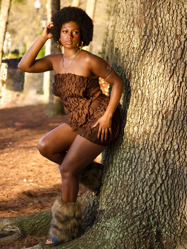 A woman in a brown dress and fur boots posing beside a tree in warm afternoon light