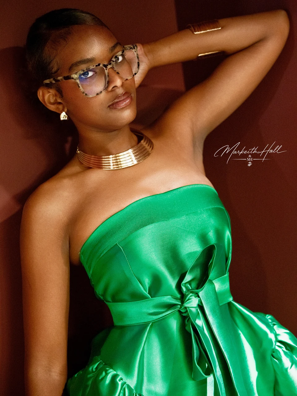 A woman in glasses and an emerald satin dress posing against a burgundy backdrop