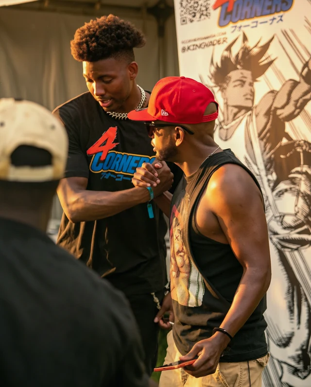 Two men clasping hands at the 4 Corners booth, one wearing a red cap